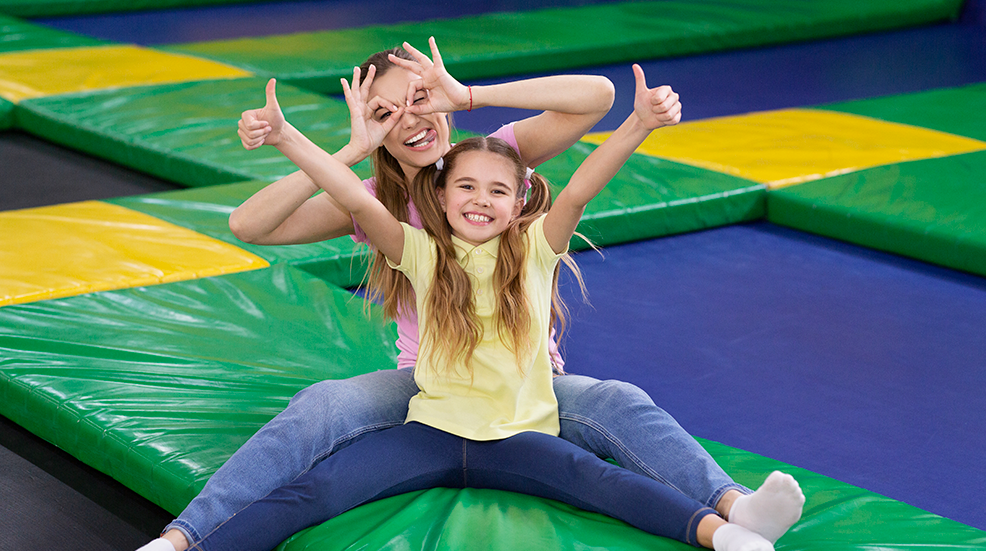 Playful silly mom with her teen daughter sitting at trampoline area of entertainment centre, showing thumbs up gesture.